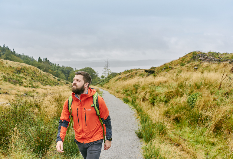 Man in orange jacket wearing haversack walking on coastal trail with loch in background, near Beinn Lora, Benderloch