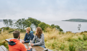 Man and two woman sit at picnic bench with views of Ardmucknish Bay in background, near Beinn Lora, Benderloch