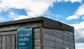 The top of a wooden wildlife hide with a blue sign saying 'Fishnish Wildlife Hide' and blue sky behind