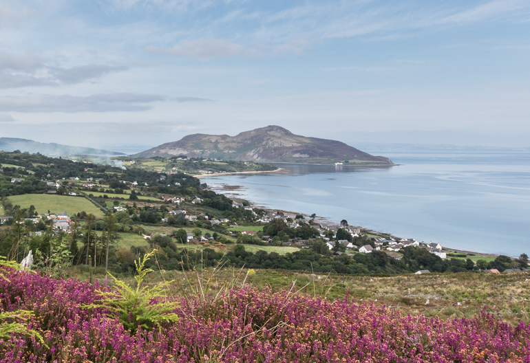 A hilltop view over heather towards fields and a hilly promontory on the coast