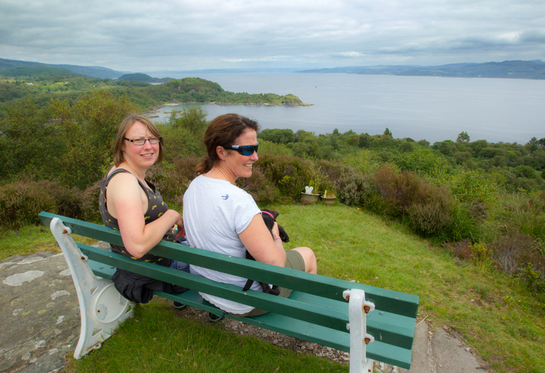 Two women sitting on a bench enjoying a view across woodland and a sea loch