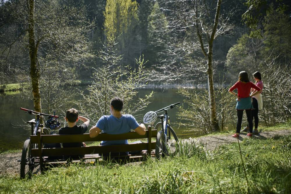 Two people sitting on a bench with bikes to either side looking out at a small pond