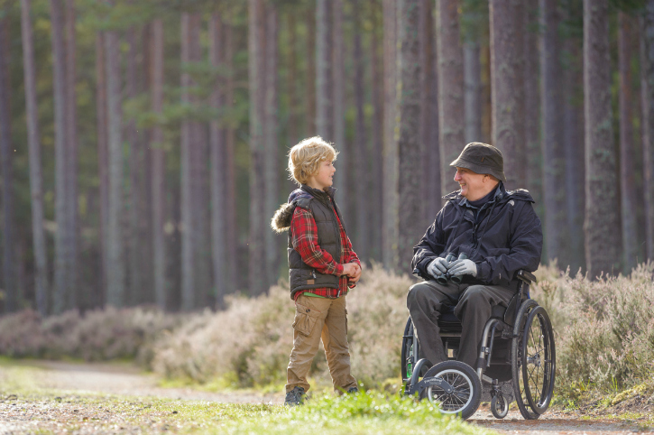 boy and man in wheelchair talk beside tall trees