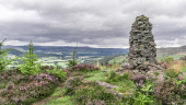 Stone carin built on the top of a hill with heather growing and a valley beyond with trees and farms