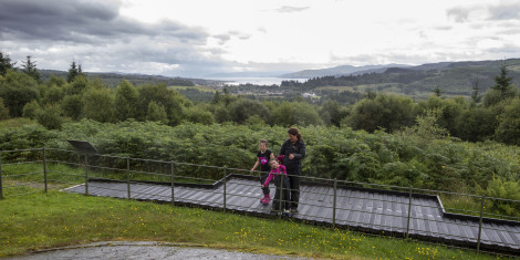 Woman and two young girls standing at railing looking at prehistoric decorated stone, with forests and loch in background, Achnabreac