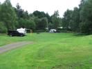 Green grassy field with gravel road through it and trees beyond, with a parked caravan