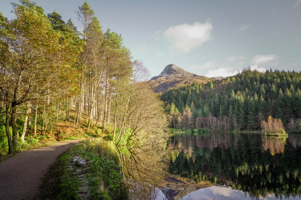 Lochan with path on the left side and forest and mountains in the background.