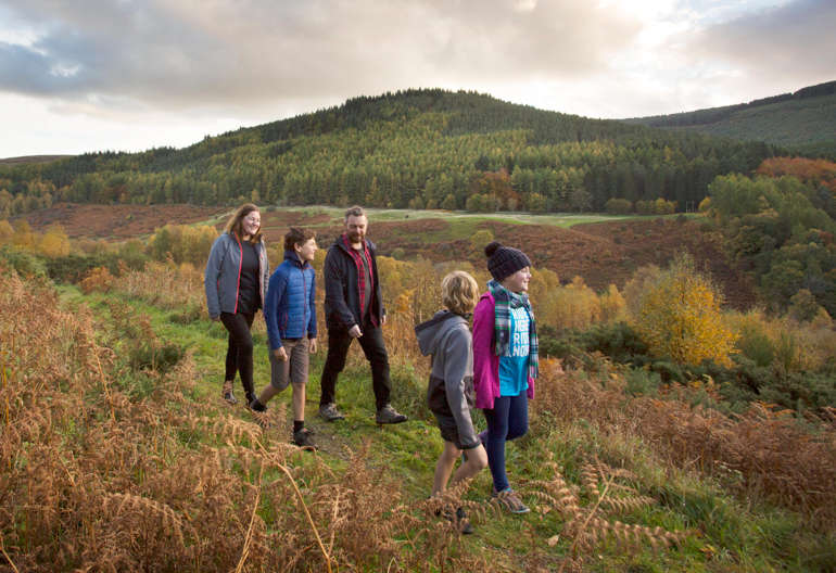 A family of 5 walk along a grassy path with views onto autumnal conifer forest on the far side of a valley