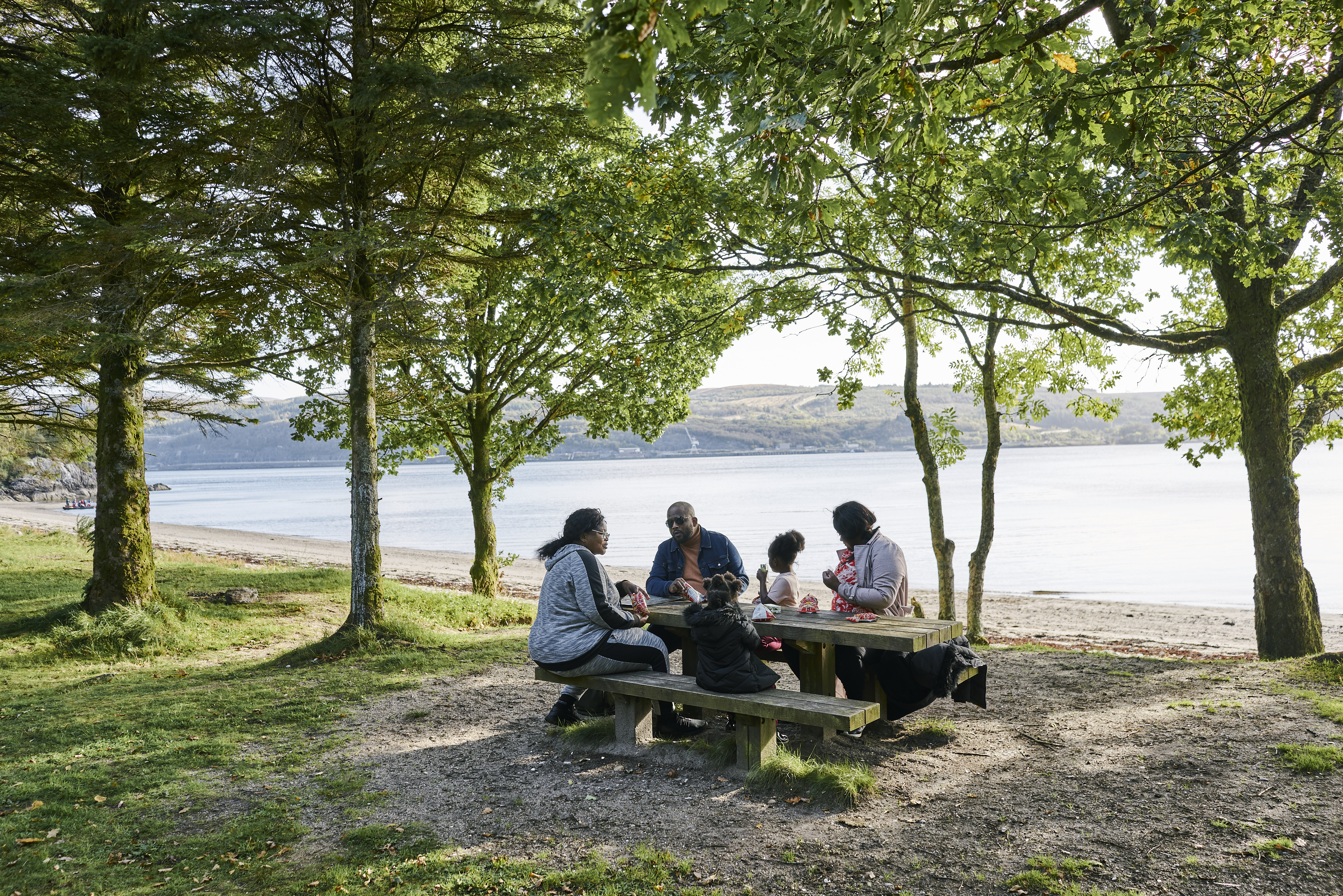 Man, two women and two young girls sit and eat at picnic table, beside beach at Ardentinny, on the shore of Loch Long