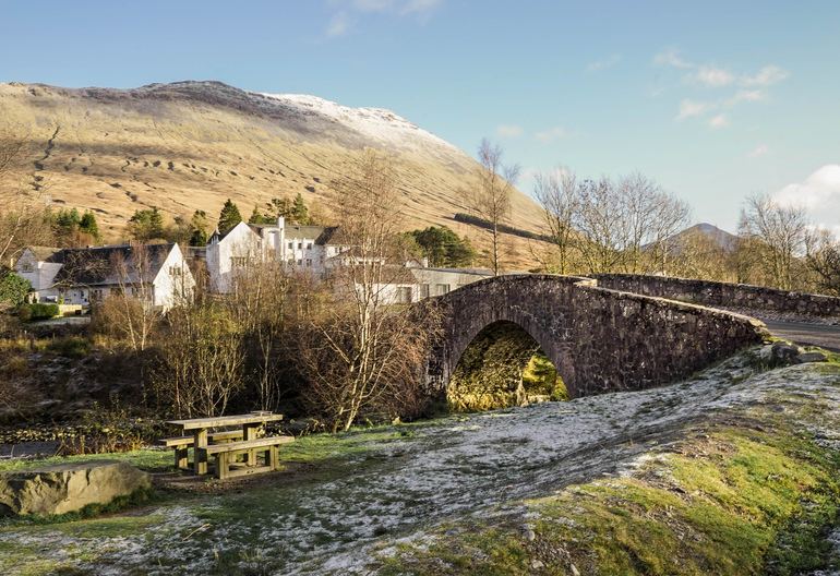 A stone bridge going towards white homes with a mountain in the background and a picnic table