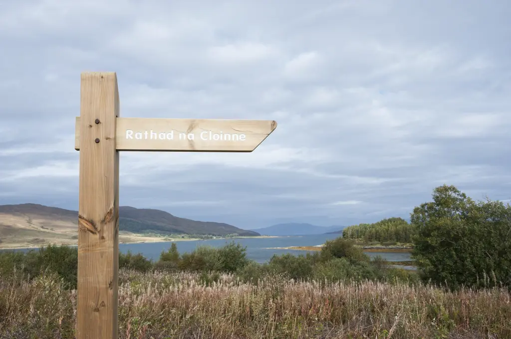Wooden sign reading Rathad na Cloinne at Broadford forest on the Isle of Skye. Wild plants, flowers and trees in the background with a view towards the sea and distant hills.