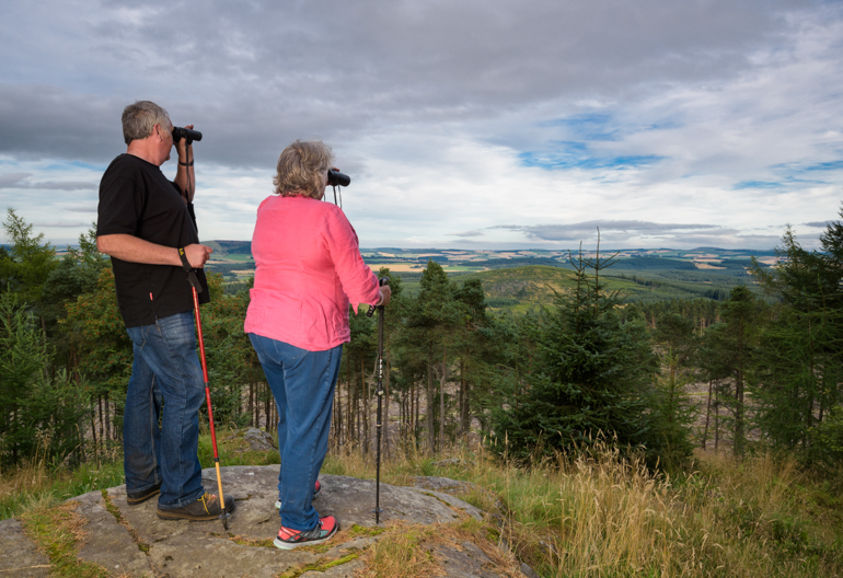 Mature male and female walker look through binoculars on vantage point, The Bin Forest, near Huntly, Aberdeenshire