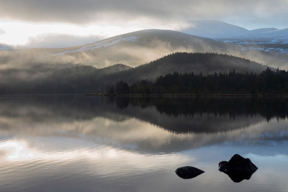 Loch in the foreground with misty forest and then snowy mountains beyond.