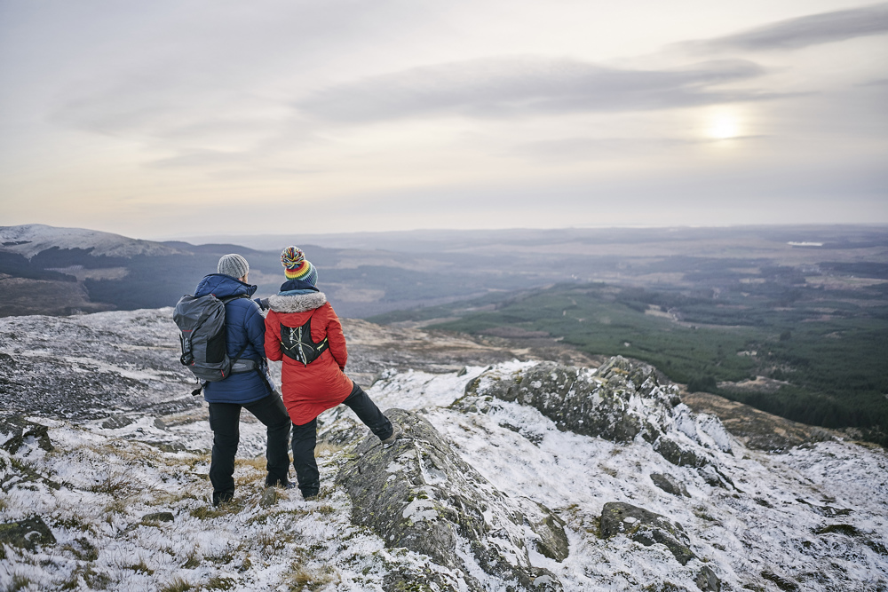 Two people on a snowy hill look over a forested landscape.