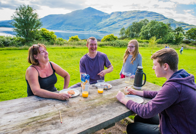 Family of four laughing while eating lunch at a picnic bench in a grassy field with loch and distant hills beyond.