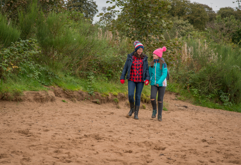 Woman and young girl walk hand in hand along the sand with woodland in background, at Boden Boo riverside beach, Renfrewshire Woods, near Erskine Bridge